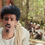 Nati Ker in a white tunic, with a coil of rope over one shoulder, singing in a forest clearing while people in white and ochre robes sit behind him: still from the official Tab Tab video