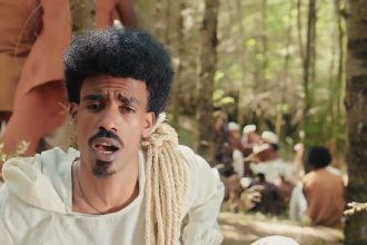 Nati Ker in a white tunic, with a coil of rope over one shoulder, singing in a forest clearing while people in white and ochre robes sit behind him: still from the official Tab Tab video