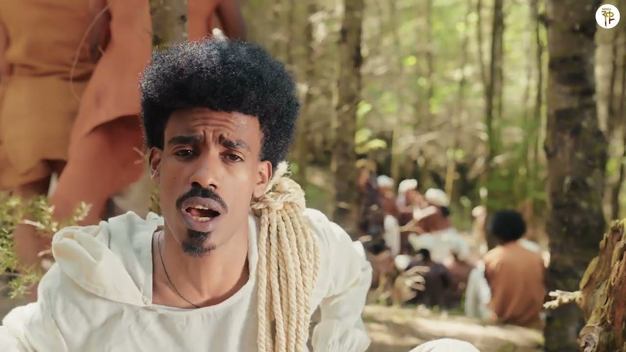 Nati Ker in a white tunic, with a coil of rope over one shoulder, singing in a forest clearing while people in white and ochre robes sit behind him: still from the official Tab Tab video
